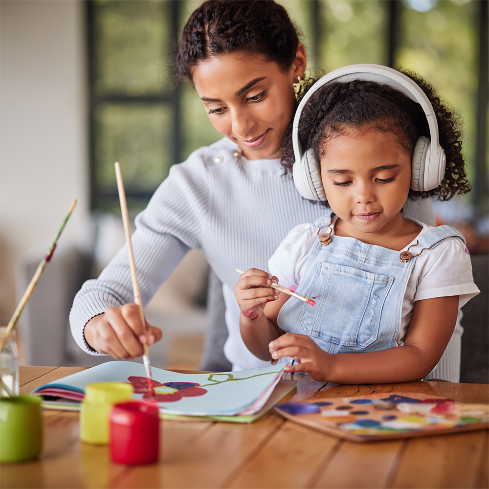 A daughter and mother painting. The daughter is on her mom's lap wearing headphones