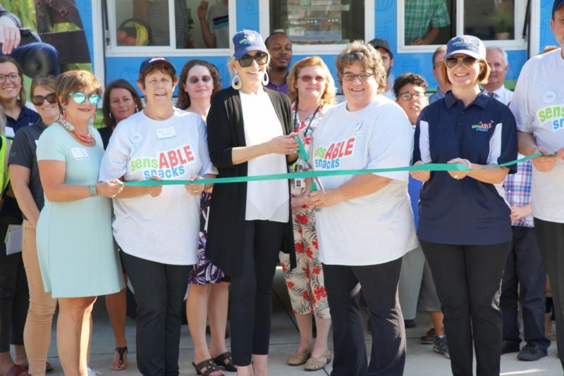 A group of Trillium and Easterseals staff hold ribbon as one person holds scissors to cut ribbon in front of Sensable Snacks food truck.